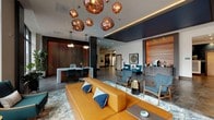 This single-floor lobby presents a thoughtfully designed contemporary space that balances visual artistry with functional comfort. The lounge area serves as the centerpiece, anchored by a substantial cognac leather sofa layered with blue accent pillows and surrounded by mid-century modern armchairs in teal and mustard tones, all arranged atop a patterned area rug. A dramatic cluster of copper globe pendant lights hangs overhead, casting a warm, sculptural focal point above the seating.

Floor-to-ceiling windows flood the lounge with natural light and offer open views to the streetscape outside. Adjacent to the lounge, a sleek white desk station with pendant lighting and dark structural columns provides a work or reception area. A striking fiber art installation — featuring colorful diagonal threads — serves as a bold decorative wall feature nearby.

The elevator lobby is finished with large-format stone-look tile flooring inlaid with multicolored mosaic accent strips, complemented by a textured stone tile accent wall and a large-scale painted artwork. Glass-paneled doors lead to additional amenity spaces, including what appears to be a fitness area. Throughout, curated artwork, warm wood accents, and a cohesive color palette of blue, copper, and natural tones create an inviting, gallery-like ambiance.