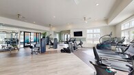 Located at 2626 Goose Island Dr in Leland, NC, this single-story fitness center is thoughtfully designed to accommodate a wide range of workout routines. The main exercise floor is expansive and open, finished with light wood-tone flooring that lends a clean, modern aesthetic. Soft sage green shiplap accent walls, recessed lighting, and multiple ceiling fans create a comfortable and inviting environment, while large windows along multiple walls flood the space with natural light and offer views of the surrounding community and pool area.

The cardio zone is well-appointed with a lineup of TRUE-brand treadmills, ellipticals, stair climbers, and stationary bikes, each equipped with personal screens. The strength training area features a comprehensive selection of cable and selectorized machines, a full dumbbell rack, and benches suited for a variety of exercises. Floor-to-ceiling mirrors expand the sense of space and support proper form during workouts.

A dedicated functional training studio adjoins the main floor, distinguished by a striking, large-scale wave mural in warm and cool tones that covers the walls. This room houses a TRX suspension training rig, a full dumbbell rack, battle ropes, and a freestanding punching bag — ideal for high-intensity and group-style training. The entry vestibule features vaulted shiplap ceilings and direct exterior access, completing this comprehensive fitness facility.