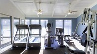 This single-story fitness space is designed to support a well-rounded cardio workout in a bright, comfortable environment. The room is outfitted with multiple treadmills, elliptical trainers, and additional cardio equipment, all arranged to maximize usability and flow throughout the space.

Architecturally, the room features an exposed beam ceiling finished in white, which adds visual interest and a sense of openness overhead. Two ceiling fans are mounted along the ceiling, promoting air circulation and maintaining a comfortable atmosphere during workouts.

A row of large windows lines the walls, flooding the interior with generous natural light and providing views of the surrounding outdoor landscape, including greenery and what appears to be a fenced exterior area. The blue-painted walls complement the bright, airy atmosphere created by the abundant sunlight.

The flooring is finished with durable, dark gym-appropriate material suited for high-traffic exercise use. Overall, the space strikes a balance between functional fitness design and an inviting, well-lit ambiance that encourages active use.