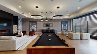 Located at 3140 E Michigan Ave in Lansing, Michigan, this contemporary amenity lounge is a thoughtfully designed social space that blends upscale aesthetics with recreational functionality. The open single-floor layout is anchored by a full-size billiards table with a sleek black felt surface, positioned beneath a striking arrangement of circular halo pendant lights and cylindrical drop pendants that illuminate the room with modern sophistication.

A dedicated lounge area features a linear electric fireplace set against a dramatic dark accent wall, accompanied by a wall-mounted display screen and a decorative log stack installation. Rich terracotta-hued barrel chairs and a plush neutral sofa surround a geometric coffee table, creating an inviting and intimate gathering spot. Large-scale black-and-white artwork adorns the textured walls throughout, adding an artistic dimension to the space.

On the opposite end, a bar counter with black seating provides a social hub, while a live-edge wood high-top table and bar chairs sit beside floor-to-ceiling windows that frame outdoor views and flood the space with natural light. The warm wood-tone flooring ties the entire room together, complementing the layered neutral and deep-toned color palette. This lounge delivers a refined, versatile environment well-suited for both relaxation and entertainment.