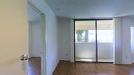 This single-floor space offers a clean, functional layout designed to maximize natural light and openness. The main living area is anchored by warm hardwood-style flooring that extends throughout, complementing the crisp white walls and textured ceiling. A standout feature of the living area is the expansive black-framed sliding glass door, which floods the space with natural light and provides direct access to an outdoor balcony. The view through the glass reveals a lush, tropical landscape with palm trees, lending the space a serene, sunlit ambiance. The open wall space offers flexibility for a variety of furnishing arrangements, while the electrical outlets are conveniently positioned throughout. A hallway visible from the main area leads to additional rooms, suggesting a thoughtfully organized floor plan. The overall aesthetic is minimal and neutral, creating a versatile canvas well-suited for a range of interior styles. The combination of natural light, tropical outdoor views, and clean finishes gives this space a bright, airy character throughout.