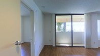 This single-floor space offers a clean, functional layout designed to maximize natural light and openness. The main living area is anchored by warm hardwood-style flooring that extends throughout, complementing the crisp white walls and textured ceiling. A standout feature of the living area is the expansive black-framed sliding glass door, which floods the space with natural light and provides direct access to an outdoor balcony. The view through the glass reveals a lush, tropical landscape with palm trees, lending the space a serene, sunlit ambiance. The open wall space offers flexibility for a variety of furnishing arrangements, while the electrical outlets are conveniently positioned throughout. A hallway visible from the main area leads to additional rooms, suggesting a thoughtfully organized floor plan. The overall aesthetic is minimal and neutral, creating a versatile canvas well-suited for a range of interior styles. The combination of natural light, tropical outdoor views, and clean finishes gives this space a bright, airy character throughout.