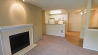 This single-floor residence offers a practical and comfortable layout designed for easy living. The main living area is anchored by a classic tile-surround fireplace with a white mantle, serving as the room's focal point. Plush neutral carpeting extends across the living space, while hardwood flooring is visible in an adjoining area, providing a warm contrast in materials underfoot.

The kitchen is positioned in an open pass-through configuration, elevated slightly above the living room and separated by a breakfast bar counter with stone-style countertops. White cabinetry lines the kitchen walls, complemented by white appliances including a refrigerator, microwave, and range, creating a clean and cohesive aesthetic. Recessed overhead lighting illuminates the kitchen workspace effectively.

The textured popcorn ceiling runs throughout the main living areas, adding a characteristic finish to the space. Warm beige and neutral tones dominate the walls and flooring, contributing to a cohesive and inviting ambiance throughout. The open sightlines between the kitchen and living areas promote a connected, functional flow, making this a well-designed single-level space ideal for comfortable everyday living.