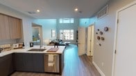 This contemporary two-story residence is designed with a clean, modern aesthetic and an open, airy floor plan. The main level centers around an open-concept kitchen and living area, where soaring double-height ceilings and large multi-pane windows flood the space with natural light and offer views of the surrounding urban landscape. The kitchen is well-appointed with light wood cabinetry, dark lower drawers, stainless steel appliances including a French door refrigerator, electric range, and built-in microwave, all complemented by sleek quartz countertops and a marble-style backsplash. A generously sized kitchen island with seating provides an ideal casual dining or gathering spot. Wood-look flooring runs consistently throughout, adding warmth to the neutral palette. The living area is anchored by large windows that frame city views. The primary bedroom features impressively tall ceilings, a dedicated workspace, and an en suite bathroom with a walk-in closet, soaking tub, and streamlined vanity. Additional bedrooms are bright and versatile, each with ample space and natural light. A full bathroom with a tub-shower combination and built-in shelving rounds out the second level. The overall ambiance is modern, bright, and urban.