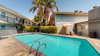 The pool area at The Solstice Apartment Homes on Casals Street in Sacramento, California, offers a well-appointed outdoor amenity space designed for relaxation and recreation. At the heart of the area is a generously sized rectangular swimming pool with clear, blue-tinted water, accessible via stainless steel entry rails. The surrounding concrete deck provides ample room for lounging, with sun chairs and a shaded seating area featuring a tan patio umbrella and wicker furnishings positioned for comfort.

A dedicated outdoor BBQ and dining station is conveniently located along one side of the deck, complete with a gas grill and patio table with chairs — ideal for outdoor gatherings. The perimeter is enclosed by a wrought iron fence with gated entry, ensuring privacy and security. Lush, mature landscaping frames the space, including a towering palm tree and dense shrubbery that provide natural shade and a tropical ambiance. The surrounding two-story residential building features balconies overlooking the pool area, enhancing the sense of community. Restroom or utility access doors are also available directly off the pool deck. Under Sacramento's abundant sunshine, this outdoor space serves as a vibrant and functional communal retreat.