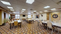 This single-floor community gathering space offers a welcoming, rustic Western ambiance defined by its bold geometric-patterned carpet in warm earth tones of gold, red, and brown that flows throughout the main areas. The centerpiece of the space is a generously sized dining and meeting room, illuminated by an array of recessed panel lighting in a drop tile ceiling. Multiple rectangular tables with padded chairs are arranged to accommodate large groups, making the room well-suited for banquets, meetings, or community events. A wall-mounted flat-screen television and a service counter along one wall add functional versatility. Natural light filters in through several windows offering views of the surrounding outdoor grounds. Adjacent to the main room, a comfortable lounge area provides a more intimate setting, furnished with plush tan sofas accented with cowhide-print pillows and a wall-mounted television. Large windows in the lounge frame outdoor views, enhancing the relaxed atmosphere. The space also includes a fully equipped kitchen featuring light maple cabinetry, granite-look countertops, black appliances including a double wall oven, range, dishwasher, refrigerator, and microwave — ideal for catering or event food preparation. Rustic decorative touches, including mounted deer antlers, framed nature artwork, and decorative accents, reinforce the cohesive Western lodge aesthetic throughout.