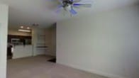 This single-floor space offers a clean and functional layout designed for comfortable everyday living. The main living area is generously sized, featuring soft carpet flooring that adds warmth underfoot and neutral-toned walls that create a bright, open atmosphere. A ceiling fan with a light fixture is centrally mounted overhead, providing both air circulation and ambient lighting throughout the room.

An open hallway or passageway connects the main living area to additional rooms beyond, offering a sense of flow and connectivity throughout the floor plan. Visible in the background are built-in shelving or cabinetry elements, suggesting organized storage solutions and thoughtful use of space. The arched or curved transition between spaces adds a subtle architectural detail that softens the overall aesthetic.

The space presents a straightforward, livable environment with a neutral palette that lends itself well to a variety of interior styles. The layout appears practical and well-proportioned, with each area transitioning smoothly into the next. Overall, this single-level space combines simple, functional design with comfortable finishes, making it a versatile and approachable living environment.