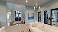 This thoughtfully designed single-floor communal space offers a refined and inviting atmosphere across several distinct lounge and gathering areas. The main entry features soaring ceilings, rich hardwood flooring, and dramatic black-framed double doors with transom windows that draw in generous natural light. A modern cylindrical chandelier adds a contemporary focal point overhead, while soft blue-gray walls lend the space a calm, sophisticated tone. Abstract artwork and clean white trim throughout reinforce a polished aesthetic.

Adjacent lounge areas transition to a cozier setting anchored by a commanding floor-to-ceiling stone fireplace with a rustic wood mantel — a standout architectural feature that adds warmth and character. Comfortable seating, including wingback chairs and plush sofas in neutral tones, creates intimate conversation areas. Black-framed French doors in this section open directly to an outdoor space, flooding the interior with natural light.

A separate gathering zone offers high ceilings, an elegant multi-arm chandelier, and a mix of seating and dining arrangements suited for both casual relaxation and group activities. Cohesive decor elements — including soft textiles, decorative accessories, and curated artwork — tie the areas together into a harmonious, welcoming environment ideal for community gatherings and social interaction.