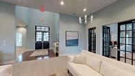 This thoughtfully designed single-floor communal space offers a refined and inviting atmosphere across several distinct lounge and gathering areas. The main entry features soaring ceilings, rich hardwood flooring, and dramatic black-framed double doors with transom windows that draw in generous natural light. A modern cylindrical chandelier adds a contemporary focal point overhead, while soft blue-gray walls lend the space a calm, sophisticated tone. Abstract artwork and clean white trim throughout reinforce a polished aesthetic.

Adjacent lounge areas transition to a cozier setting anchored by a commanding floor-to-ceiling stone fireplace with a rustic wood mantel — a standout architectural feature that adds warmth and character. Comfortable seating, including wingback chairs and plush sofas in neutral tones, creates intimate conversation areas. Black-framed French doors in this section open directly to an outdoor space, flooding the interior with natural light.

A separate gathering zone offers high ceilings, an elegant multi-arm chandelier, and a mix of seating and dining arrangements suited for both casual relaxation and group activities. Cohesive decor elements — including soft textiles, decorative accessories, and curated artwork — tie the areas together into a harmonious, welcoming environment ideal for community gatherings and social interaction.