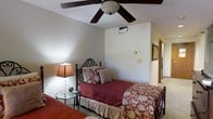 This single-floor space presents a well-appointed bedroom designed with comfort and classic style in mind. The room features neutral-toned walls that create a calm, inviting atmosphere, complemented by a ceiling fan with dark wood-toned blades that provides both air circulation and a traditional aesthetic touch. A wall-mounted HVAC vent ensures climate control year-round.

The sleeping area accommodates multiple beds, including a twin or full bed with an elegant wrought iron headboard adorned with scrollwork detailing, dressed in rich burgundy bedding with decorative accent pillows. A second bed with a wooden frame and matching floral-patterned bedding is partially visible in the foreground, suggesting ample sleeping capacity.

A small circular glass-topped side table sits between sleeping areas, holding a warm-toned table lamp with a square shade and a crystal-style base, adding a soft ambient glow to the room. Framed artwork is visible along the adjoining hallway wall, adding a decorative touch. An open doorway leads to an adjacent space, hinting at a well-connected layout. The overall ambiance is warm, traditional, and functional.