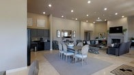This single-floor space showcases an open-concept layout that seamlessly integrates a dining area, lounge seating, and a kitchenette or wet bar into one cohesive environment. The soaring ceilings amplify the sense of spaciousness, while recessed lighting provides consistent, warm illumination throughout the interior.

The dining area serves as a central focal point, anchored by a substantial rectangular table surrounded by elegantly upholstered French-style chairs in a neutral gray tone, set atop a large area rug. A decorative mirror and framed artwork above a sideboard add visual depth to this zone.

Adjacent to the dining area, a comfortable lounge section features a generously sized sectional sofa with accent pillows, accompanied by a stylish coffee table, creating an inviting space for relaxed gatherings.

Along one wall, a dedicated wet bar or kitchenette is outfitted with dark cabinetry, under-counter appliances, and a coffee station, offering practical amenities for entertaining. Light-toned tile flooring runs throughout, complementing the neutral beige walls and contributing to the overall contemporary, refined ambiance of the space.