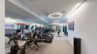This fitness center is a thoughtfully designed, single-floor facility offering a comprehensive range of equipment and training zones. The space is organized into two primary areas: a cardio zone and a strength and functional training zone, connected by an open, flowing layout that maximizes usability.

The cardio zone features a generous row of elliptical trainers, stationary spin bikes, and a rowing machine, all positioned along a full-length mirrored wall that visually expands the space. Circular halo-style pendant lights are suspended from the white drop ceiling, providing bright, even illumination throughout. Neutral carpet tiles cover the floor, contributing to a clean, professional aesthetic.

The strength and functional training zone is anchored by a striking red-and-black Octagon multi-functional rig, equipped with a pull-up bar, cable attachments, and extensive free weight storage including dumbbells, kettlebells, plates, and battle ropes. A heavy punching bag adds a boxing element, while resistance bands and exercise mats support flexibility and bodyweight training. A wall-mounted flat-screen television and a water station further enhance the facility's functionality. Large windows in the strength area allow natural light to enter, offering views of surrounding greenery.