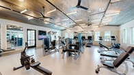 Located at 8830 College Pkwy in Fort Myers, FL, this fitness center offers a spacious, single-floor layout designed to support a comprehensive workout experience. The space is defined by its standout architectural feature: a warm, wood-plank ceiling accented by exposed black metal rods, pendant-style linear lighting fixtures, and ceiling fans that contribute both to the aesthetic appeal and air circulation throughout the facility.

The main workout floor is finished with a light-colored, easy-to-clean surface that spans the entire area. Large wall mirrors line key sections of the room, enhancing the sense of openness and allowing users to monitor their form during exercise. Multiple windows along the walls invite natural light, creating a bright and energizing atmosphere.

The cardio zone is well-appointed with a row of treadmills and additional aerobic machines, each equipped with personal screens. The strength training area features a comprehensive selection of Life Fitness cable and selectorized machines, free weight benches, a squat rack, and rowing machines to accommodate a full range of training needs.

A dedicated functional training zone includes a pull-up rig and open floor space for bodyweight or stretching exercises. Built-in shelving, a cushioned bench, water fountains, and a wall-mounted television round out the amenities, making this fitness center both functional and comfortable for daily use.