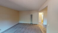Located at 964 Mangrove Ave in Sunnyvale, California, this single-story unit offers a straightforward and functional layout across several interconnected rooms. The living area is generously sized, finished with wall-to-wall carpet and neutral-toned walls beneath a textured ceiling. A wall-mounted heater unit provides climate control, and a large sliding window brings in natural light while offering a view of the outdoor greenery and fencing. The bedroom is similarly carpeted and bright, featuring its own window and a ceiling fan for added comfort, with a wall heater also present. The galley-style kitchen is well-appointed with rich, warm-toned wood cabinetry running floor to ceiling on both sides, white laminate countertops, a double-basin sink, and tile flooring. A window at the end of the kitchen provides natural light and a pleasant outdoor view. White appliances, including a refrigerator and range, complete the kitchen. The bathroom features tile flooring, a wide vanity with a single-basin sink, a large framed mirror with overhead lighting, and ample under-sink storage. The overall layout, as visible in the 3D overview, is compact yet efficient, with rooms flowing naturally from one to the next in a cohesive, single-level arrangement.