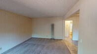Located at 964 Mangrove Ave in Sunnyvale, California, this single-story unit offers a straightforward and functional layout across several interconnected rooms. The living area is generously sized, finished with wall-to-wall carpet and neutral-toned walls beneath a textured ceiling. A wall-mounted heater unit provides climate control, and a large sliding window brings in natural light while offering a view of the outdoor greenery and fencing. The bedroom is similarly carpeted and bright, featuring its own window and a ceiling fan for added comfort, with a wall heater also present. The galley-style kitchen is well-appointed with rich, warm-toned wood cabinetry running floor to ceiling on both sides, white laminate countertops, a double-basin sink, and tile flooring. A window at the end of the kitchen provides natural light and a pleasant outdoor view. White appliances, including a refrigerator and range, complete the kitchen. The bathroom features tile flooring, a wide vanity with a single-basin sink, a large framed mirror with overhead lighting, and ample under-sink storage. The overall layout, as visible in the 3D overview, is compact yet efficient, with rooms flowing naturally from one to the next in a cohesive, single-level arrangement.