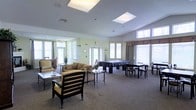 This single-floor common area is designed to accommodate a variety of activities and social interactions. The space is generously sized, featuring multiple skylights in the vaulted ceiling that flood the interior with natural light, complemented by several large windows along the perimeter walls. The soft, neutral color palette—with light green walls and warm carpeting—creates a welcoming and relaxed atmosphere throughout.

The layout is thoughtfully divided into distinct functional zones. A comfortable lounge area anchors one section of the room, furnished with a plush sofa, cushioned armchairs, and a glass-topped coffee table, encouraging casual conversation and relaxation. Adjacent to this is a games area centered around a billiards table, providing a focal point for recreational use. A dining or activity table with chairs occupies another corner, offering space for seated gatherings or group activities.

Natural light is a defining feature, with windows dressed in layered curtains and valances that allow for flexible light control. The open floor plan and absence of interior walls ensure a seamless flow between zones, making the space adaptable for both small gatherings and larger community events.