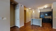 This single-floor residence offers a thoughtfully designed open-concept layout that seamlessly connects the living and kitchen areas. Rich, dark hardwood-style flooring runs throughout, adding warmth and a sense of continuity to the space. The modern kitchen is a standout feature, anchored by a sleek peninsula-style island with a light stone countertop that provides both prep space and casual seating. Pendant lighting hangs above the island, complementing the track lighting nearby to create a well-lit, inviting atmosphere. Dark cabinetry in the kitchen adds a bold contrast to the lighter walls and countertops, while built-in appliances contribute to the clean, streamlined aesthetic. The open hallway beyond the main living area leads to multiple doors, suggesting well-organized access to additional rooms and storage spaces. Tall ceilings enhance the sense of openness, and HVAC vents are thoughtfully integrated into the upper walls to maintain comfort throughout. Neutral-toned walls provide a versatile backdrop, making the space easy to personalize. Overall, the residence balances modern finishes with a functional, open layout suited for everyday living.