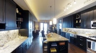 This elegant single-floor residence showcases a thoughtfully designed open-concept layout that seamlessly connects the kitchen, dining, and living areas into one cohesive living space. The kitchen is a standout feature, anchored by a generous island topped with striking light-veined granite, offering bar seating and built-in shelving below. Rich, dark cabinetry lines the walls from floor to ceiling, lending a bold, sophisticated contrast to the lighter countertops and backsplash. Under-cabinet lighting adds a warm, functional glow, while pendant lights and a track lighting system overhead ensure the space is well-illuminated throughout. Dark hardwood flooring extends across the entire open floor plan, creating a continuous sense of depth and warmth. The kitchen flows naturally into a dining area and then into a cozy living room, where large glass doors allow natural light to filter in and provide a glimpse of an outdoor balcony or terrace beyond. The living area is furnished with inviting seating and ambient lighting, completing the warm and refined atmosphere. The overall aesthetic is modern yet welcoming, with a careful balance of dark tones and warm lighting that gives the entire space a polished, upscale character.