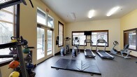 This fitness center offers a bright and functional environment designed to support a well-rounded workout routine. Large glass doors and multiple windows flood the space with natural light, providing views of the outdoors and creating an open, energizing atmosphere. The warm yellow walls and light-toned ceiling contribute to an inviting and motivating setting.

The cardio area is well-appointed with multiple treadmills and an elliptical trainer, all positioned to take advantage of the natural light and outdoor views. A flat-screen television is mounted on the wall, offering entertainment during workouts. The flooring features a durable, rubberized surface in the equipment zones, while padded exercise mats are laid out in a dedicated stretching and floor exercise area, providing ample space for yoga, stretching, or bodyweight exercises.

Additional fitness equipment, including free weights and a stationary bike, is available along the perimeter of the room. The space is neatly organized to maximize usability without feeling cluttered. Direct access to an outdoor area is provided through the glass double doors, which open to what appears to be a pool or exterior recreation area, seamlessly connecting indoor fitness activities with outdoor amenities.