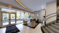 This sophisticated hotel lobby makes a striking first impression with its refined architectural details and carefully curated design elements. The space is defined by a sweeping curved ceiling adorned with warm recessed cove lighting, which adds a soft, welcoming glow throughout the area. Large floor-to-ceiling windows and glass entry doors frame views of the streetscape beyond, bathing the interior in generous natural light and creating a seamless connection to the outdoors. The flooring features polished light-toned tiles that enhance the sense of openness, accented by a bold branded entrance mat. A central seating arrangement anchors the space, composed of patterned accent chairs and a dark sofa set atop a geometric rug, offering a comfortable and stylish area for guests to gather. The walls are dressed in an ornate, metallic-toned damask wallcovering that lends the space a sense of luxury and grandeur. Decorative pendant lanterns and wall sconces complement the ambient lighting scheme, while a potted plant adds a natural, organic touch. A staircase with a sleek railing is visible to one side, indicating vertical connectivity within the building. Overall, the lobby exudes a polished and inviting atmosphere, balancing classic elegance with contemporary comfort.