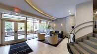 This sophisticated hotel lobby makes a striking first impression with its refined architectural details and carefully curated design elements. The space is defined by a sweeping curved ceiling adorned with warm recessed cove lighting, which adds a soft, welcoming glow throughout the area. Large floor-to-ceiling windows and glass entry doors frame views of the streetscape beyond, bathing the interior in generous natural light and creating a seamless connection to the outdoors. The flooring features polished light-toned tiles that enhance the sense of openness, accented by a bold branded entrance mat. A central seating arrangement anchors the space, composed of patterned accent chairs and a dark sofa set atop a geometric rug, offering a comfortable and stylish area for guests to gather. The walls are dressed in an ornate, metallic-toned damask wallcovering that lends the space a sense of luxury and grandeur. Decorative pendant lanterns and wall sconces complement the ambient lighting scheme, while a potted plant adds a natural, organic touch. A staircase with a sleek railing is visible to one side, indicating vertical connectivity within the building. Overall, the lobby exudes a polished and inviting atmosphere, balancing classic elegance with contemporary comfort.