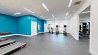 This spacious fitness center offers a comprehensive workout environment across a single, open floor plan. The interior design features a striking combination of bold teal accent walls contrasted against crisp white walls and ceilings, creating an energizing atmosphere for exercise. Durable rubber flooring in a speckled gray finish covers the entire space, providing safety and comfort underfoot throughout all workout zones.

The facility is divided into distinct functional areas. The cardio zone is equipped with multiple treadmills, stationary bikes, and elliptical machines, all positioned near large windows that allow natural light to flow in and offer views of the surrounding greenery. A dedicated strength training area houses a cable machine, selectorized resistance equipment, and a Sportwall reaction training system mounted to the teal accent wall.

A separate section provides space for functional training and flexibility, featuring a heavy punching bag, stability balls, and a large wall mirror to assist with form. Wall-mounted flat-screen TVs are positioned throughout to provide entertainment during workouts.

Illumination is provided by rows of modern linear LED fixtures mounted flush to the white ceilings, ensuring bright, even lighting across the entire facility. Multiple windows and glass-paneled entry doors further enhance the sense of openness, connecting the interior to the outdoors while maintaining a clean, modern aesthetic.