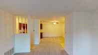 This single-floor space presents a functional and straightforward layout designed for comfortable everyday living. The main living area is generously sized, featuring neutral-toned carpet that extends throughout the space, creating a cohesive and understated aesthetic. A partial dividing wall with decorative spindles separates the entry area from the main living space, adding a subtle architectural detail while maintaining an open feel. A ceiling fan overhead provides both light and air circulation to the adjoining room.

The living area connects seamlessly to a secondary space, which benefits from a sliding glass door dressed with vertical blinds, allowing natural light to filter in while providing access to the outdoors. An additional carpeted room features a large grid-pane window that frames an exterior view of greenery, filling the space with natural light.

The kitchen is equipped with warm wood-toned cabinetry offering ample storage, light-colored countertops, a double-basin sink, a dishwasher, an electric range, a built-in microwave, and a refrigerator. Tile flooring in the kitchen provides a practical transition from the carpeted living areas.

The bathroom includes a combined tub and shower unit with sliding glass doors, a toilet, vanity storage, and tile flooring, offering a clean and efficient design throughout.