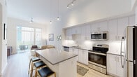 Located at 1710 N Congress Ave in West Palm Beach, FL, this thoughtfully designed two-bedroom, one-bathroom residence offers a clean, contemporary aesthetic throughout its efficiently organized single floor. The open-concept kitchen and living area serve as the heart of the home, highlighted by dramatically high ceilings and pendant lighting that draw the eye upward. The kitchen is outfitted with white shaker-style cabinetry, a white subway tile backsplash, stainless steel appliances including a refrigerator, electric range, microwave, and dishwasher, and a generous quartz-topped island with bar seating — ideal for casual dining or entertaining. Light wood-look flooring runs throughout, lending warmth and continuity. The living area is bathed in natural light from large sliding glass doors with vertical blinds that open to an outdoor view, creating a seamless indoor-outdoor feel. The primary bedroom is a comfortable retreat featuring a large window with outdoor views, while the second bedroom offers additional flexibility. The bathroom includes a white vanity with quartz countertop, a framed mirror, and a tub/shower combination with marble-style tile surround. A hallway connects all spaces efficiently, completing this well-appointed West Palm Beach residence.