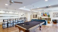 This single-floor community lounge and recreation space offers a thoughtfully designed, open-concept layout that seamlessly blends entertainment, relaxation, and social dining. The generously sized main area is anchored by a full-size billiards table and a shuffleboard table, providing ample room for recreation without feeling crowded. Light hardwood flooring runs throughout, complementing the bright, white walls and recessed ceiling lighting that give the space a clean, modern aesthetic. A coffered ceiling detail adds architectural interest and helps define zones within the open floor plan.

The lounge area features a plush blue sectional sofa, accent chairs, a wall-mounted large-screen TV, and a built-in shelving unit — creating a comfortable space for relaxing or gathering. A large-format Scrabble board mounted on the wall adds a playful, decorative touch. An exposed brick wall and a glass garage-style door flood the space with natural light and lend an urban, industrial character.

The kitchen area is fully equipped with stainless steel appliances, quartz countertops, white cabinetry, and a striking tiled accent wall featuring green, white, and copper mosaic detailing. Pendant lighting and a bar-height counter with seating complete the space, making it well-suited for hosting events and social gatherings.