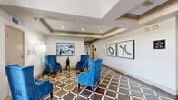 Located at 2727 Elmside Dr in Houston, TX, this single-floor community space offers a generously sized, thoughtfully designed interior that balances elegance with functionality. A grand entrance lobby greets visitors with a soaring arched glass facade that floods the space with natural light, complemented by a decorative lantern pendant and a multi-story ceiling that creates a dramatic first impression. The distinctive multicolored slate tile flooring, laid in a bold checkerboard pattern, flows seamlessly throughout the entire space, providing visual continuity and a refined aesthetic. The main lounge area features recessed lighting, crown molding, decorative column details, and a built-in fireplace niche, with ample seating arranged around dual arched mirrors. An additional seating alcove offers a cozy built-in bench framed by plantation shutters and vibrant accent artwork, bathed in natural light from large windows. A glass-walled private office or meeting room provides transparency while maintaining acoustic separation. The kitchen is efficiently appointed with white quartz countertops, stainless steel appliances including a refrigerator and dishwasher, and a pass-through opening to the lounge. Built-in bookshelves and accent navy walls add character and warmth throughout, making this a versatile and well-equipped gathering space.