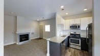 This single-floor residence showcases a thoughtfully designed open-concept layout that seamlessly connects the kitchen and living areas. The kitchen is outfitted with stainless steel appliances, including a gas range and built-in microwave, complemented by crisp white cabinetry and a classic white subway tile backsplash. Dark charcoal lower cabinets provide a striking contrast beneath the light quartz countertops, which extend into a peninsula with an integrated sink — ideal for casual dining or meal preparation. Wood-look flooring anchors the kitchen, transitioning into plush carpet throughout the living area. The living space is highlighted by a sleek, modern fireplace set against a clean white surround, adding warmth and a focal point to the room. Recessed lighting illuminates the space throughout, contributing to a bright and contemporary atmosphere. Neutral gray walls and white trim create a cohesive, modern aesthetic. A entry door with a sidelight window is visible in the background, allowing natural light to filter into the space.