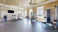 Located at 8585 Woodway Dr in Houston, TX, this fitness center offers a well-appointed, single-level exercise space designed to support a full range of workout routines. The room features notably high ceilings that enhance the sense of openness, while multiple large, navy-framed windows along one wall flood the interior with abundant natural light and provide views of the surrounding greenery. Three ceiling fans are suspended overhead, promoting consistent airflow throughout the space.

The cardio section is well-stocked with treadmills, elliptical trainers, stationary bikes, and a rowing machine, all positioned to take advantage of the natural light from the windows. A multi-station cable machine anchors the strength-training area, complemented by a dumbbell rack, adjustable benches, and additional cable attachment equipment mounted along the walls. Wall-mounted flat-screen televisions are positioned on opposing walls for convenient viewing during workouts.

Neutral-toned carpet tiles cover the flooring throughout, while large framed mirrors on one wall visually expand the space and allow users to monitor their form. Navy blue accent trim along the baseboards and door frames ties the design together, giving the facility a clean, modern aesthetic. Blue-framed glass doors provide a transparent entry, maintaining an open and inviting atmosphere.