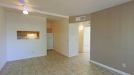 This single-floor residence presents a straightforward and functional layout designed for comfortable everyday living. The main living area is finished with light-toned ceramic tile flooring throughout, contributing to a clean, easy-to-maintain environment. Neutral beige and white wall tones keep the space feeling bright and open.

The kitchen is positioned adjacent to the living area and features a convenient pass-through window, allowing for easy interaction between the two spaces. Wood-toned cabinetry and a white refrigerator are visible, providing practical storage and appliance space. Overhead ventilation ensures proper airflow throughout.

A central wall partition separates the living area from a hallway that leads to additional rooms, offering a sense of defined zones within the open plan. A wall-mounted thermostat is visible, indicating centralized climate control. Ceiling vents are well-placed to ensure consistent air distribution across the unit.

The hallway beyond the partition is illuminated by a wall sconce, lending a warm transition to the adjacent rooms. White baseboards frame each room cleanly, adding a crisp finish to the neutral color palette. Overall, the space conveys a practical and tidy residential environment suited for functional, day-to-day living.