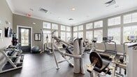 This fitness center is a thoughtfully designed, single-floor facility that offers a comprehensive workout environment for users of all fitness levels. The space is defined by its warm gray walls, white crown molding, and durable rubber flooring, creating a clean and professional atmosphere conducive to exercise.

Large windows with adjustable blinds line multiple walls, flooding the interior with natural light and providing views of the surrounding outdoor greenery. Recessed ceiling lighting supplements the natural light, ensuring a well-lit environment throughout the day.

The cardio zone is generously equipped with multiple treadmills, elliptical trainers, and stationary bikes—primarily from True Fitness—arranged to allow for comfortable spacing between users. The strength-training area features a cable and pulley multi-station machine, a leg press, a leg extension/curl machine, a weight bench with rack, and a dedicated free weights section with a dumbbell rack.

Additional amenities include stability balls, wall-mounted fitness instructional artwork, a water fountain station, and accessible exits. The layout is open and efficient, maximizing usable floor space while maintaining clear pathways throughout. The overall ambiance is bright, functional, and well-maintained, making this fitness center a practical and inviting space for daily exercise.