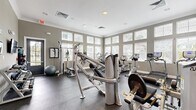 This fitness center is a thoughtfully designed, single-floor facility that offers a comprehensive workout environment for users of all fitness levels. The space is defined by its warm gray walls, white crown molding, and durable rubber flooring, creating a clean and professional atmosphere conducive to exercise.

Large windows with adjustable blinds line multiple walls, flooding the interior with natural light and providing views of the surrounding outdoor greenery. Recessed ceiling lighting supplements the natural light, ensuring a well-lit environment throughout the day.

The cardio zone is generously equipped with multiple treadmills, elliptical trainers, and stationary bikes—primarily from True Fitness—arranged to allow for comfortable spacing between users. The strength-training area features a cable and pulley multi-station machine, a leg press, a leg extension/curl machine, a weight bench with rack, and a dedicated free weights section with a dumbbell rack.

Additional amenities include stability balls, wall-mounted fitness instructional artwork, a water fountain station, and accessible exits. The layout is open and efficient, maximizing usable floor space while maintaining clear pathways throughout. The overall ambiance is bright, functional, and well-maintained, making this fitness center a practical and inviting space for daily exercise.