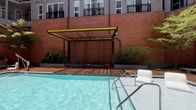 Located at 401 W Harnett Street in downtown Raleigh, this single-level outdoor courtyard is part of The Dillon, a mixed-use development distinguished by its industrial-chic aesthetic and urban energy. The space is framed by warm brick facades and expansive floor-to-ceiling glass walls, allowing visual connectivity between the interior and exterior environments.

The courtyard is anchored by a resort-style swimming pool, bordered by a wood-decked cabana structure featuring a slatted pergola with signature yellow steel accents and cushioned lounge seating. String lights overhead add an inviting ambiance for evening use.

The main gathering area features generously spaced seating clusters with wood-framed upholstered chairs arranged around coffee tables, shaded by mature deciduous trees that provide natural canopy throughout the paved courtyard. Bold yellow bar-height tables and stools offer casual seating options nearby.

An outdoor kitchen equipped with a stainless steel grill provides amenities for entertaining. A covered outdoor lounge area houses a mounted flat-screen television alongside a built-in grill station. Sculptural woven chaise lounges add artistic character to the paved walkways, complemented by a large-scale mosaic mural on the brick wall depicting a swimmer, reinforcing the space's playful, design-forward identity.
