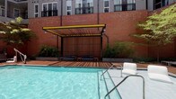 Located at 401 W Harnett Street in downtown Raleigh, this single-level outdoor courtyard is part of The Dillon, a mixed-use development distinguished by its industrial-chic aesthetic and urban energy. The space is framed by warm brick facades and expansive floor-to-ceiling glass walls, allowing visual connectivity between the interior and exterior environments.

The courtyard is anchored by a resort-style swimming pool, bordered by a wood-decked cabana structure featuring a slatted pergola with signature yellow steel accents and cushioned lounge seating. String lights overhead add an inviting ambiance for evening use.

The main gathering area features generously spaced seating clusters with wood-framed upholstered chairs arranged around coffee tables, shaded by mature deciduous trees that provide natural canopy throughout the paved courtyard. Bold yellow bar-height tables and stools offer casual seating options nearby.

An outdoor kitchen equipped with a stainless steel grill provides amenities for entertaining. A covered outdoor lounge area houses a mounted flat-screen television alongside a built-in grill station. Sculptural woven chaise lounges add artistic character to the paved walkways, complemented by a large-scale mosaic mural on the brick wall depicting a swimmer, reinforcing the space's playful, design-forward identity.