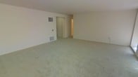 This single-floor space presents a generously sized main living area finished with wall-to-wall carpeting in a neutral tone, creating a clean and understated foundation suitable for a variety of interior styles. The walls are painted in a soft, light color that contributes to an open and airy feel throughout the room. A wall-mounted HVAC vent and a floor-level return air grille are visibly integrated into one wall, indicating a central climate control system. A doorway visible toward the rear of the living area leads to an adjoining hallway or secondary space, suggesting additional rooms beyond the main area. The layout appears straightforward and functional, with smooth transitions between spaces. The ceiling height is standard, keeping the atmosphere comfortable and well-proportioned. Overall, the space offers a blank canvas with practical built-in systems, making it well-suited for customization to meet a range of living needs.