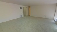 This single-floor space presents a generously sized main living area finished with wall-to-wall carpeting in a neutral tone, creating a clean and understated foundation suitable for a variety of interior styles. The walls are painted in a soft, light color that contributes to an open and airy feel throughout the room. A wall-mounted HVAC vent and a floor-level return air grille are visibly integrated into one wall, indicating a central climate control system. A doorway visible toward the rear of the living area leads to an adjoining hallway or secondary space, suggesting additional rooms beyond the main area. The layout appears straightforward and functional, with smooth transitions between spaces. The ceiling height is standard, keeping the atmosphere comfortable and well-proportioned. Overall, the space offers a blank canvas with practical built-in systems, making it well-suited for customization to meet a range of living needs.