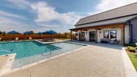 Located at 705 E Bluebird Dr in Lafayette, Louisiana, this single-level outdoor amenity space is designed for relaxation and community gathering. At its centerpiece is a generously sized, crystal-clear swimming pool with a connected shallow-entry spa, both finished with a clean concrete surround that offers ample space for lounging. Surrounding the pool are multiple chaise lounges shaded by vibrant blue market umbrellas, providing comfortable spots to soak up the Louisiana sun. A covered patio with exposed wood-beam construction and French doors connects the indoor clubhouse to the pool deck, offering shaded seating and a seamless indoor-outdoor flow. Adjacent to the pool area, a beautifully landscaped courtyard features lush artificial turf, manicured garden beds with fresh mulch, young trees, and a central fire pit surrounded by Adirondack chairs. Overhead string lights crisscross the courtyard, creating an inviting atmosphere for evening gatherings. Dining tables with umbrellas provide additional outdoor seating. The entire area is enclosed by a warm-toned wood privacy fence, giving the space a sense of seclusion while maintaining an open, airy feel under expansive Louisiana skies.
