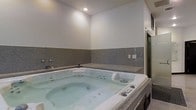 This single-floor space showcases a dedicated indoor spa area centered around a generously sized jetted jacuzzi tub. The tub is framed by a granite tile surround that extends along the lower portion of the walls, adding a polished, upscale aesthetic to the room. The upper walls are finished in a clean, neutral tone, creating a calm and relaxing ambiance throughout. Recessed ceiling lighting provides soft, focused illumination, enhancing the spa-like atmosphere. The jacuzzi itself features multiple jets and built-in controls, designed for a premium hydrotherapy experience. A granite ledge runs along the back wall, offering a convenient surface for toiletries or décor. A wall-mounted faucet is visible to the side, and an adjacent doorway leads to what appears to be a separate shower or additional bathroom area, adding to the overall functionality of the space. The combination of granite finishes, neutral tones, and thoughtful lighting creates an environment well-suited for relaxation and rejuvenation.