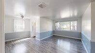 Located at 1313 E San Miguel St in Colorado Springs, CO, this single-floor unit presents a clean, well-maintained interior with a cohesive design aesthetic throughout. The most prominent design feature is the two-tone wall treatment found across multiple rooms — a soft, muted blue on the lower half paired with crisp white above — creating a calm and consistent visual identity throughout the space.

The main living area is generously proportioned, featuring durable wood-style laminate flooring in a gray tone that complements the color palette. Multiple windows fitted with vertical blinds allow abundant natural light to fill the room, offering views of the surrounding greenery. A ceiling fan is present in the living area, and a central HVAC vent is visible overhead. The layout flows openly toward what appears to be a kitchen area, where a white refrigerator is visible.

Additional rooms continue the two-tone wall design and wood-style flooring. One room includes a closet with a clothing rod, suitable for use as a bedroom. The bathroom features a full tub and shower combination enclosed by classic white square tile. The kitchen includes an electric range and a window overlooking outdoor greenery, allowing natural light into the cooking area. Overall, the space offers a tidy, functional layout with consistent finishes throughout.