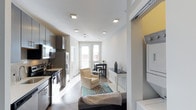 This single-floor space is designed with an open-concept layout that seamlessly connects the kitchen, living, and dining areas in one continuous flow. The kitchen, positioned along the left wall, is outfitted with stainless steel appliances, dark cabinetry, and ample counter space, providing a functional and modern cooking environment. Recessed lighting throughout the ceiling ensures consistent illumination across the entire space.

The living area features hardwood-style flooring and is furnished with a neutral-toned armchair and sofa, anchored by a patterned area rug that adds warmth and definition to the zone. Natural light pours in through large double doors at the far end of the space, which also offer an exterior view, creating a bright and airy ambiance throughout.

Adjacent to the living area, a compact dining space accommodates a small table and chairs, making efficient use of the available footprint. A notable practical feature is the dedicated in-unit laundry area, visible on the right side, which houses a stacked washer and dryer unit tucked neatly behind a sliding door. The overall aesthetic is clean, contemporary, and functional.
