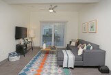 This 4-bedroom, 2-bathroom unit is thoughtfully designed across a single floor, offering a functional and modern living environment well-suited for shared occupancy. The open-concept common area seamlessly integrates the kitchen and living space, creating a communal hub that feels both spacious and inviting. The kitchen is outfitted with stainless steel appliances, including a full-size refrigerator, and features a clean two-tone cabinet design with white upper cabinets and dark lower cabinets, complemented by light wood-look luxury vinyl plank flooring that flows throughout the space. Recessed lighting and a ceiling fan enhance both comfort and illumination in the living area, which includes room for a sofa, coffee table, and entertainment setup. A sliding glass door with vertical blinds allows natural light to fill the space while providing outdoor access. Each bedroom is comfortably sized to accommodate a full or queen bed, a desk, and personal storage. The bedrooms feature vertical blind-covered windows or sliding glass doors that invite natural light, with neutral walls that offer a versatile backdrop. The overall ambiance is clean, contemporary, and practical, designed to support a comfortable day-to-day lifestyle.