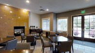 This single-floor hospitality space offers a warm and functional dining environment designed for comfort and convenience. The room is anchored by a prominent floor-to-ceiling fireplace clad in golden-toned tile, serving as a striking focal point that adds character and warmth to the overall ambiance. The space is furnished with multiple dark wood dining tables and upholstered chairs in neutral tones, providing ample seating for guests across several distinct arrangements. A self-serve beverage station, complete with coffee urns and a dark wood credenza, is thoughtfully positioned near the fireplace wall, offering easy access for visitors. Large windows dressed with white horizontal blinds allow generous natural light to fill the room, offering views of the surrounding greenery outside. The walls are finished in a contemporary two-tone palette of warm gray and white, complementing the light tile flooring underfoot. An exterior door provides direct access to the outdoors, enhancing the sense of openness. The layout balances an intimate lounge corner — featuring patterned accent chairs — with more traditional dining seating, making the space versatile for both casual gatherings and relaxed individual use.