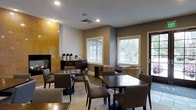 This single-floor hospitality space offers a warm and functional dining environment designed for comfort and convenience. The room is anchored by a prominent floor-to-ceiling fireplace clad in golden-toned tile, serving as a striking focal point that adds character and warmth to the overall ambiance. The space is furnished with multiple dark wood dining tables and upholstered chairs in neutral tones, providing ample seating for guests across several distinct arrangements. A self-serve beverage station, complete with coffee urns and a dark wood credenza, is thoughtfully positioned near the fireplace wall, offering easy access for visitors. Large windows dressed with white horizontal blinds allow generous natural light to fill the room, offering views of the surrounding greenery outside. The walls are finished in a contemporary two-tone palette of warm gray and white, complementing the light tile flooring underfoot. An exterior door provides direct access to the outdoors, enhancing the sense of openness. The layout balances an intimate lounge corner — featuring patterned accent chairs — with more traditional dining seating, making the space versatile for both casual gatherings and relaxed individual use.