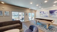 This well-appointed game room in San Antonio, Texas, offers a versatile and inviting amenity space designed for both leisure and social interaction. The room is finished with crown molding, recessed lighting, and a soft blue-toned ceiling that lends a polished, contemporary feel throughout.

At the heart of the space is a billiards table set atop a decorative area rug, accompanied by plush lounge seating including a tufted sofa and rounded accent chairs upholstered in cool blue tones. Abstract wall art adds a artistic touch to the neutral-toned walls. Large sliding glass doors and generous windows flood the room with natural light while offering direct views of the outdoor pool and patio area, creating a seamless indoor-outdoor connection.

A dedicated dining and activity zone features multiple round wooden tables paired with modern chairs, alongside tall dark-wood storage cabinetry. A wall-mounted flat-screen television is positioned for comfortable viewing from the seating area, making it suitable for watching events or gaming.

The flooring is covered with patterned carpeting that ties together the room's cohesive design scheme. The overall ambiance is modern, comfortable, and community-oriented — well-suited for casual gatherings, recreation, and socializing.