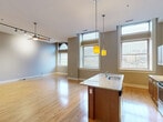 Located at 1800 Fifth Ave in Pittsburgh's Fifth Avenue corridor, this single-floor unit offers a thoughtfully designed open-concept layout that seamlessly integrates the living, dining, and kitchen areas into one expansive, light-filled space. Polished hardwood floors flow throughout, reflecting the generous natural light that pours in through large, tall windows — including a charming arched window accent. Soaring ceilings amplify the sense of space, while neutral gray walls create a clean, modern backdrop.

The kitchen is a standout feature, anchored by a substantial granite-topped island with an integrated sink and pendant lighting overhead. Warm-toned wood cabinetry pairs with stainless steel appliances, including a range, microwave, and refrigerator, offering both style and practicality. The living area is equipped with ceiling fan and track lighting for versatile illumination.

The bathroom features tile flooring, a dark-finished vanity with a light countertop, and well-appointed fixtures. Two closets provide ample storage, while a dedicated laundry area adds everyday convenience. The unit's well-considered layout balances open, social spaces with functional private areas, making it a practical and attractive home in the heart of Pittsburgh.