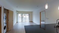 This single-floor space presents a clean, modern aesthetic with an emphasis on open living. The main living area is generously proportioned, featuring light hardwood flooring that extends throughout and smooth, white walls that enhance the sense of brightness and space. Large sliding glass doors flood the room with natural light and open to a private balcony, offering views of the surrounding urban streetscape. A wall-mounted split air conditioning unit provides climate control, seamlessly integrated into the room's minimalist design. Warm wood-toned door frames add a subtle contrast to the crisp white walls, lending a contemporary yet inviting character to the interior. A doorway with matching wood trim leads to an adjacent room, which is finished with carpeted flooring — offering a softer, quieter environment suitable for a bedroom or study. The overall ambiance is airy and uncluttered, with a neutral color palette that provides a versatile canvas for a variety of interior styles. The thoughtful layout and abundance of natural light make this a highly functional and comfortable living space.