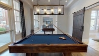 This well-appointed game room is designed to deliver an engaging recreational experience, combining multiple entertainment zones within a single, cohesive space. The centerpiece is a full-size billiards table with a navy felt surface and a rich walnut finish, positioned beneath a striking rectangular chandelier fitted with industrial-style glass cylinder pendants that cast warm, focused light over the playing surface. A foosball table nearby provides an additional layer of competitive fun. The room flows seamlessly into a lounge area featuring an octagonal poker table surrounded by stylish spotted upholstered chairs. This corner is anchored by a stone-clad fireplace with built-in cabinetry on either side and a wall-mounted flat-screen television above, creating an inviting entertainment nook. Large windows throughout the space allow generous natural light to pour in, offering views of the surrounding exterior. Rich hardwood flooring extends across the entire room, complementing the warm, contemporary design aesthetic. A distinctive reclaimed wood sliding barn door adds a rustic-chic accent and serves as a functional room divider. Decorative wall art, including starburst mirrors and abstract canvas pieces, contribute to the polished, thoughtfully curated atmosphere of this versatile gathering space.